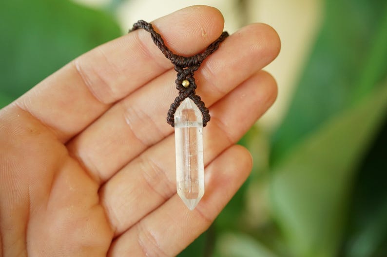 May include: A clear, double-terminated quartz crystal pendant secured in a dark brown macrame cord setting. The necklace features a small, round, metallic gold-colored bead accent positioned directly above the crystal point in the woven cord.