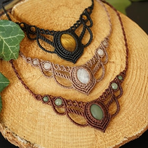 May include: Three macrame necklaces with gemstone pendants rest on a wooden surface. The top necklace is black with a tiger's eye stone, the middle is tan with a white stone, and the bottom is brown with a green stone.