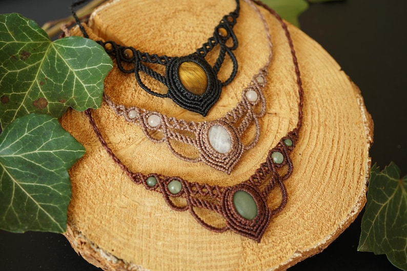 May include: Three macrame necklaces with gemstone pendants rest on a wooden surface. The top necklace is black with a tiger's eye stone, the middle is tan with a white stone, and the bottom is brown with a green stone.