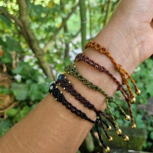 May include: A collection of macrame bracelets in various earth-tone colors, including black, brown, green, and gold. Each bracelet features a unique woven design and is adorned with small gold beads and adjustable ties. The bracelets are displayed on a wrist.
