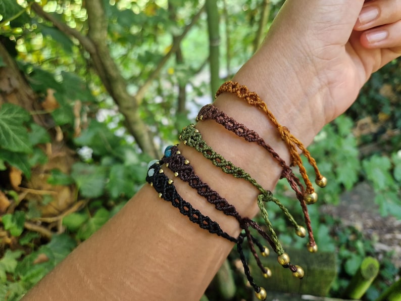 May include: A collection of macrame bracelets in various earth-tone colors, including black, brown, green, and gold. Each bracelet features a unique woven design and is adorned with small gold beads and adjustable ties. The bracelets are displayed on a wrist.