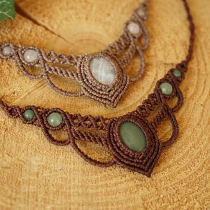 May include: Two handmade macrame necklaces with gemstone accents. One necklace features a brown cord with green stones, while the other has a lighter brown cord with white stones. The necklaces are displayed on a wooden surface.