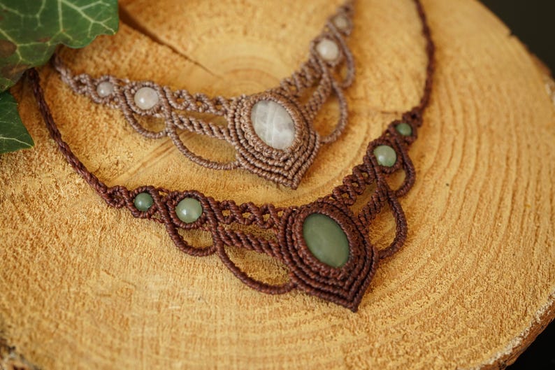 May include: Two handmade macrame necklaces with gemstone accents. One necklace features a brown cord with green stones, while the other has a lighter brown cord with white stones. The necklaces are displayed on a wooden surface.