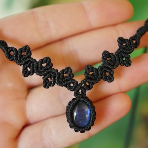 Puede incluir: Un collar de macram&eacute; negro con una piedra preciosa azul de forma ovalada engastada en un cord&oacute;n anudado. La banda del collar consiste en una serie de patrones de nudos decorativos y sim&eacute;tricos que se extienden desde el colgante central.
