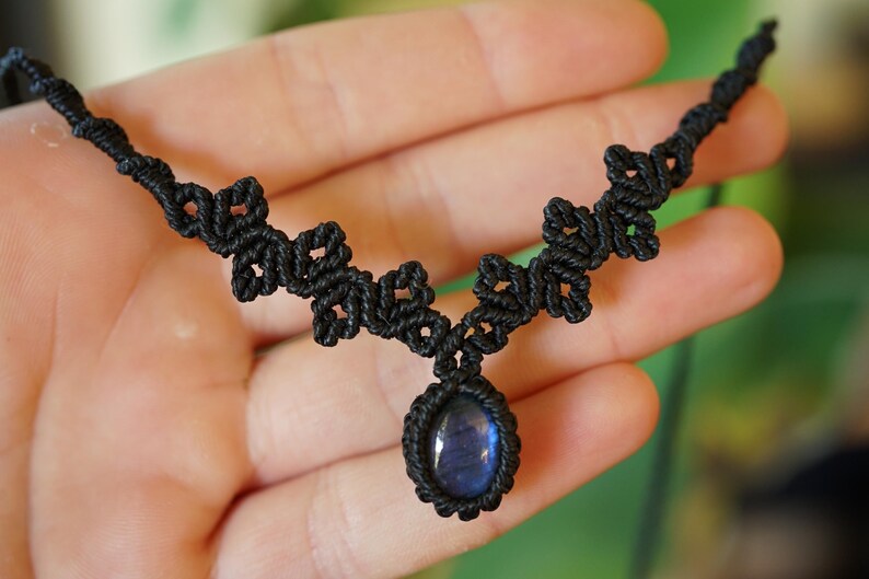 Puede incluir: Un collar de macram&eacute; negro con una piedra preciosa azul de forma ovalada engastada en un cord&oacute;n anudado. La banda del collar consiste en una serie de patrones de nudos decorativos y sim&eacute;tricos que se extienden desde el colgante central.