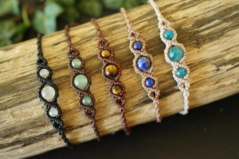 May include: Five macrame bracelets with gemstone beads in various colors, including white, green, brown, yellow, blue, and turquoise. The bracelets are displayed on a piece of wood, showcasing the intricate knotting and natural stone accents.