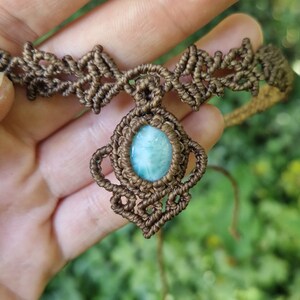 May include: A handcrafted macrame necklace featuring a central oval-shaped, light blue gemstone pendant. The necklace is made with brown cord, showcasing intricate knotting patterns. The background is blurred, with green foliage.