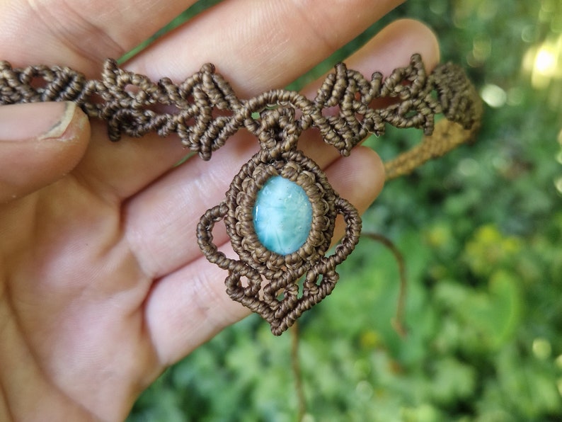 May include: A handcrafted macrame necklace featuring a central oval-shaped, light blue gemstone pendant. The necklace is made with brown cord, showcasing intricate knotting patterns. The background is blurred, with green foliage.