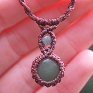 May include: A handmade macrame necklace featuring a central green gemstone pendant and a smaller green bead. The necklace is crafted with brown cord, showcasing intricate knotting details. The design is simple and elegant.