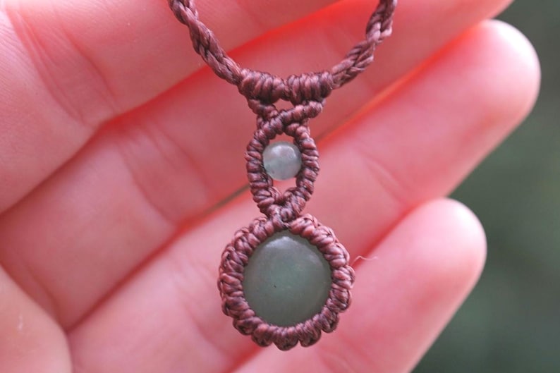 May include: A handmade macrame necklace featuring a central green gemstone pendant and a smaller green bead. The necklace is crafted with brown cord, showcasing intricate knotting details. The design is simple and elegant.