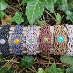 May include: A collection of macrame bracelets with various colored gemstones, including gray, yellow, green, brown, turquoise, and purple. The bracelets are displayed on a piece of wood, surrounded by green leaves.