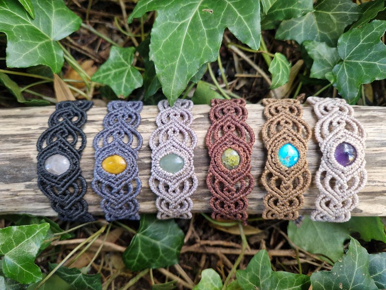 May include: A collection of macrame bracelets with various colored gemstones, including gray, yellow, green, brown, turquoise, and purple. The bracelets are displayed on a piece of wood, surrounded by green leaves.
