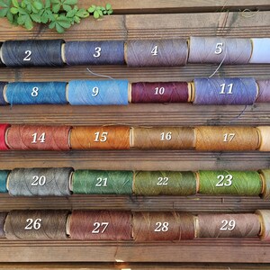 May include: Thirty cylindrical spools of thread arranged in five rows of six, each labeled with a white number from 1 to 30. The spools display a diverse color palette including black, gray, white, blue, red, brown, green, and various earth tones.