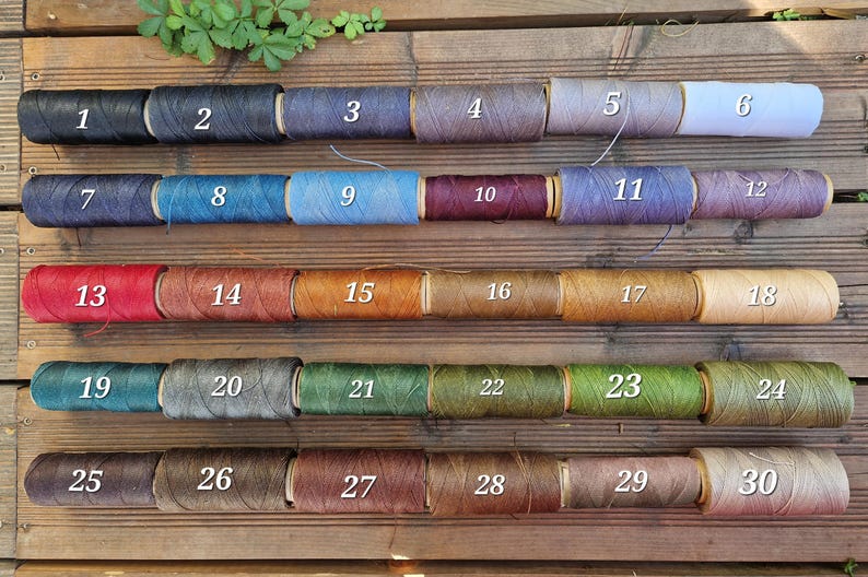 May include: Thirty cylindrical spools of thread arranged in five rows of six, each labeled with a white number from 1 to 30. The spools display a diverse color palette including black, gray, white, blue, red, brown, green, and various earth tones.