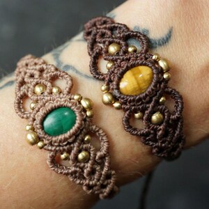 Könnte beinhalten: Zwei Makramee-Armbänder mit komplexen geknoteten Kordelmustern in Hellbraun und Dunkelbraun. Jedes Armband ist mit einem ovalen Edelstein besetzt, einem grünen Malachit und einem goldenen Tigerauge, akzentuiert durch kleine metallische Perlen.