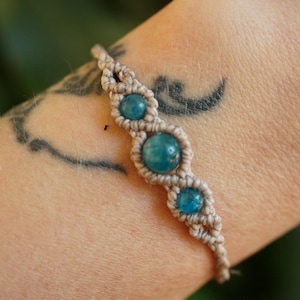 May include: A macrame bracelet featuring three round, turquoise-colored beads. The bracelet is made with beige-colored cord and is worn on a person's arm. The arm has a tattoo on it.