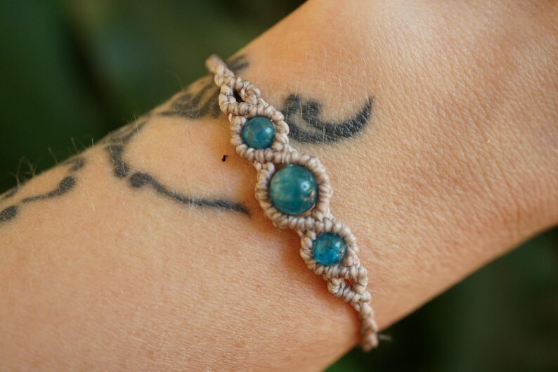 May include: A macrame bracelet featuring three round, turquoise-colored beads. The bracelet is made with beige-colored cord and is worn on a person's arm. The arm has a tattoo on it.