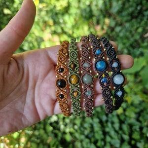 May include: Five macrame bracelets with gemstone beads in various colors, including brown, green, and black. Each bracelet features a unique gemstone, such as tiger's eye and aquamarine, and is accented with small gold beads.