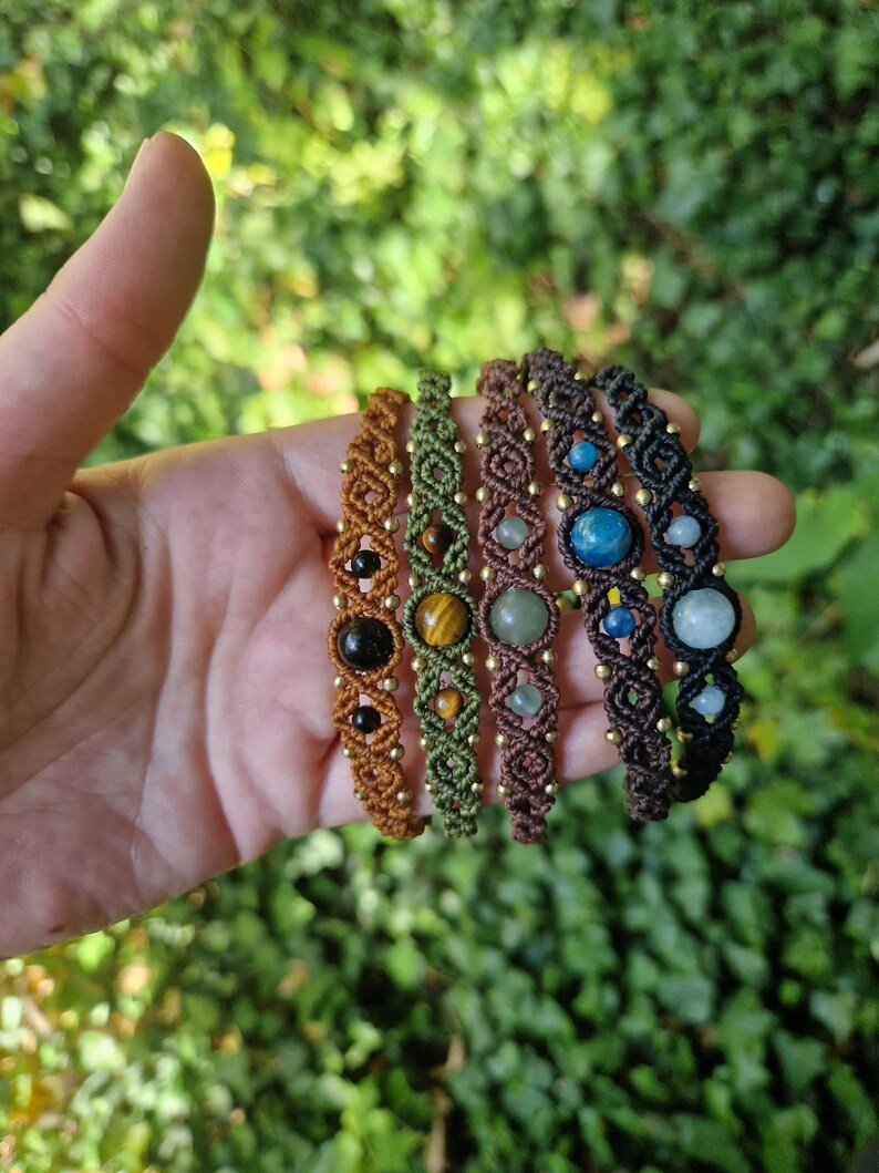 May include: Five macrame bracelets with gemstone beads in various colors, including brown, green, and black. Each bracelet features a unique gemstone, such as tiger's eye and aquamarine, and is accented with small gold beads.