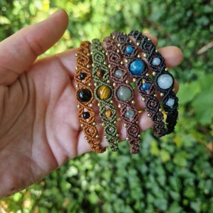 May include: A hand displays a collection of macrame bracelets. Each bracelet features a different gemstone, including black onyx, tiger's eye, and blue apatite. The bracelets are in shades of brown, green, and black, with gold-colored beads.