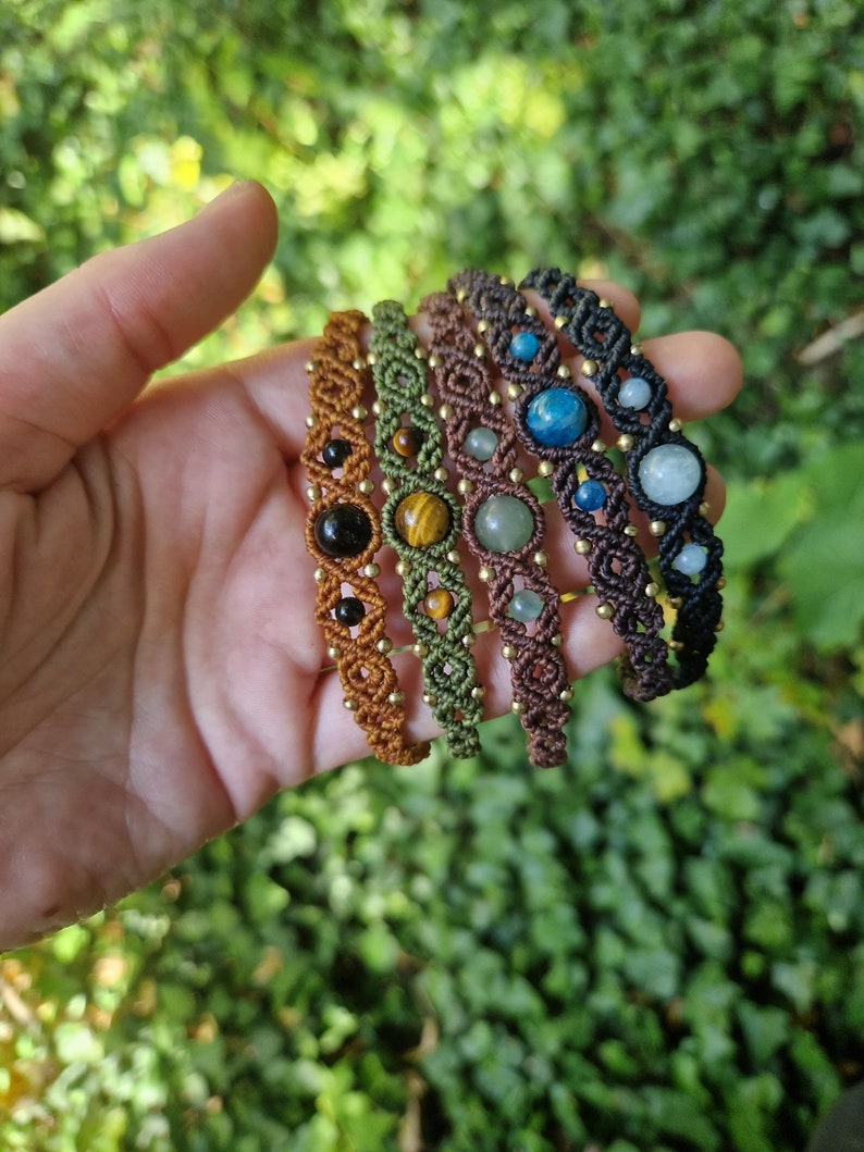 May include: A hand displays a collection of macrame bracelets. Each bracelet features a different gemstone, including black onyx, tiger's eye, and blue apatite. The bracelets are in shades of brown, green, and black, with gold-colored beads.