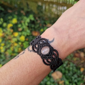 May include: Black macrame bracelet featuring a central oval rose quartz stone. The bracelet is intricately knotted and worn on a wrist. The background is blurred with green and yellow foliage.