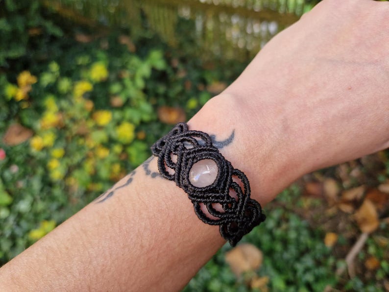 May include: Black macrame bracelet featuring a central oval rose quartz stone. The bracelet is intricately knotted and worn on a wrist. The background is blurred with green and yellow foliage.
