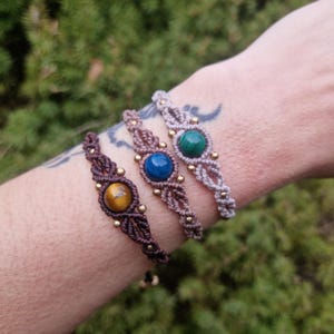 May include: Three macrame bracelets with gemstone centerpieces are displayed. The bracelets feature brown, blue, and green stones, accented with small gold beads. The bracelets are on a wrist, with a blurred green background.