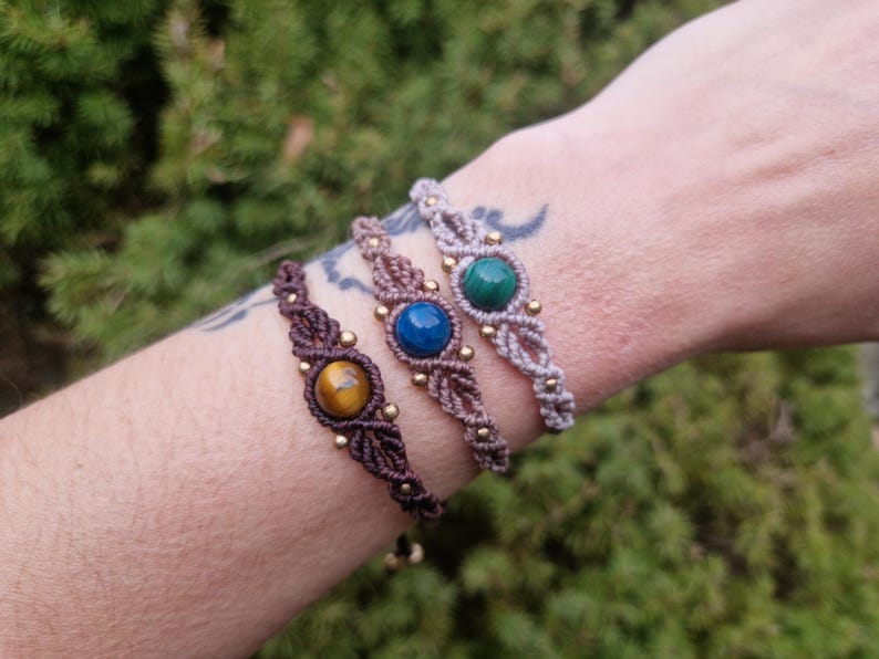 May include: Three macrame bracelets with gemstone centerpieces are displayed. The bracelets feature brown, blue, and green stones, accented with small gold beads. The bracelets are on a wrist, with a blurred green background.