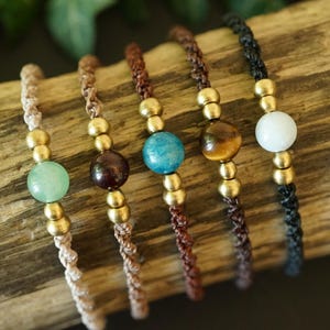 May include: Five macrame bracelets with gold-colored beads and gemstone accents. The gemstones are green, burgundy, blue, brown, and white. The bracelets are displayed on a piece of wood.