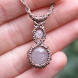 May include: A handmade macrame necklace featuring a light pink gemstone pendant. The necklace has a braided, tan-colored cord with a small gemstone above the larger pendant. The necklace is held in a hand.