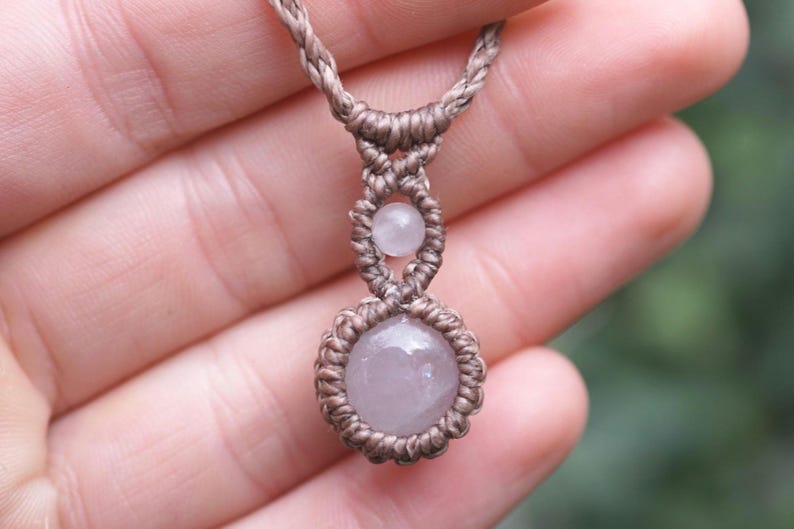 May include: A handmade macrame necklace featuring a light pink gemstone pendant. The necklace has a braided, tan-colored cord with a small gemstone above the larger pendant. The necklace is held in a hand.