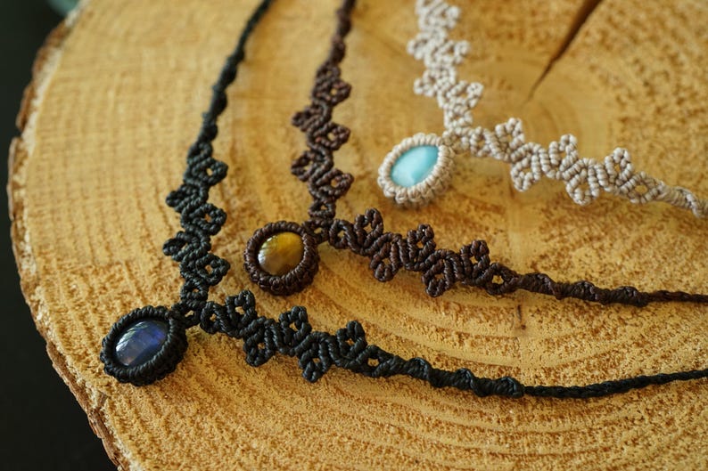 Puede incluir: Tres collares de macram&eacute; con intrincados patrones de cord&oacute;n anudado. Un collar es negro con una piedra ovalada azul oscuro, otro es marr&oacute;n oscuro con una piedra ojo de tigre dorada y otro es beige con una piedra ovalada azul claro.
