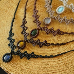 Puede incluir: Una colecci&oacute;n de cuatro collares de macram&eacute; con dise&ntilde;os de cord&oacute;n anudado en negro, marr&oacute;n oscuro y beige. Cada collar est&aacute; centrado con una piedra preciosa ovalada, incluyendo piedras azules, ojo de tigre, verde oscuro y azul claro.