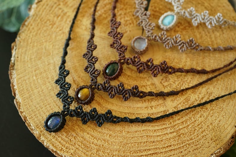 Puede incluir: Una colecci&oacute;n de cuatro collares de macram&eacute; con dise&ntilde;os de cord&oacute;n anudado en negro, marr&oacute;n oscuro y beige. Cada collar est&aacute; centrado con una piedra preciosa ovalada, incluyendo piedras azules, ojo de tigre, verde oscuro y azul claro.
