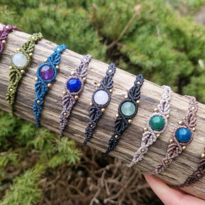 May include: A collection of macrame bracelets with various gemstone centerpieces. The bracelets feature a woven design in colors like purple, green, blue, gray, and brown, with gold-colored beads. The gemstones include amethyst, jade, and tiger's eye.