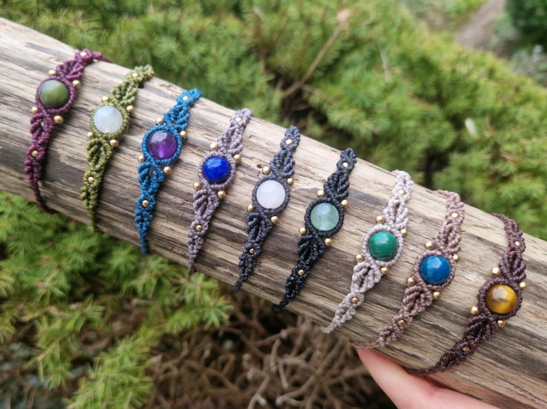 May include: A collection of macrame bracelets with various gemstone centerpieces. The bracelets feature a woven design in colors like purple, green, blue, gray, and brown, with gold-colored beads. The gemstones include amethyst, jade, and tiger's eye.