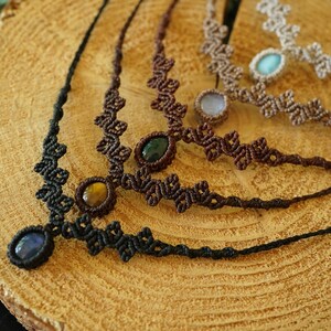 Puede incluir: Una colecci&oacute;n de cuatro collares de macram&eacute; con intrincados patrones de cord&oacute;n anudado en negro, marr&oacute;n oscuro y beige. Cada collar est&aacute; centrado con una piedra preciosa ovalada pulida, incluyendo piedras azules, ojo de tigre, verdes y azul claro.