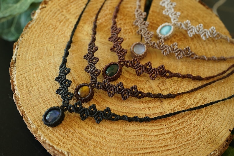 Puede incluir: Una colecci&oacute;n de cuatro collares de macram&eacute; con intrincados patrones de cord&oacute;n anudado en negro, marr&oacute;n oscuro y beige. Cada collar est&aacute; centrado con una piedra preciosa ovalada pulida, incluyendo piedras azules, ojo de tigre, verdes y azul claro.