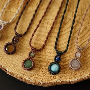 May include: A collection of handmade macrame necklaces with gemstone pendants. The necklaces feature various colors of cord and different gemstones, including amethyst, jade, and rose quartz. The necklaces are displayed on a wooden surface.