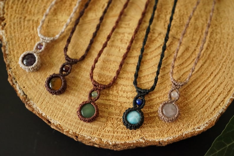 May include: A collection of handmade macrame necklaces with gemstone pendants. The necklaces feature various colors of cord and different gemstones, including amethyst, jade, and rose quartz. The necklaces are displayed on a wooden surface.