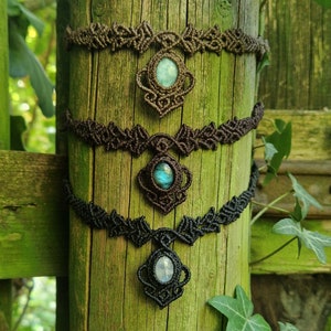 May include: Three macrame chokers in varying shades of brown and black, each featuring a central gemstone pendant. The gemstones are in shades of blue and green. The necklaces are displayed on a weathered wooden post.