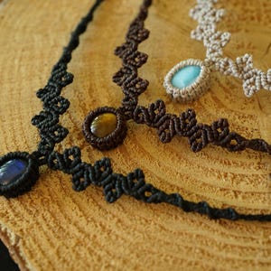 Puede incluir: Tres collares de macram&eacute; con piedras cabuj&oacute;n ovaladas. Un collar es negro con una piedra azul oscuro, otro es marr&oacute;n oscuro con una piedra ojo de tigre marr&oacute;n dorado y otro es beige con una piedra azul claro, todos con nudos intrincados.