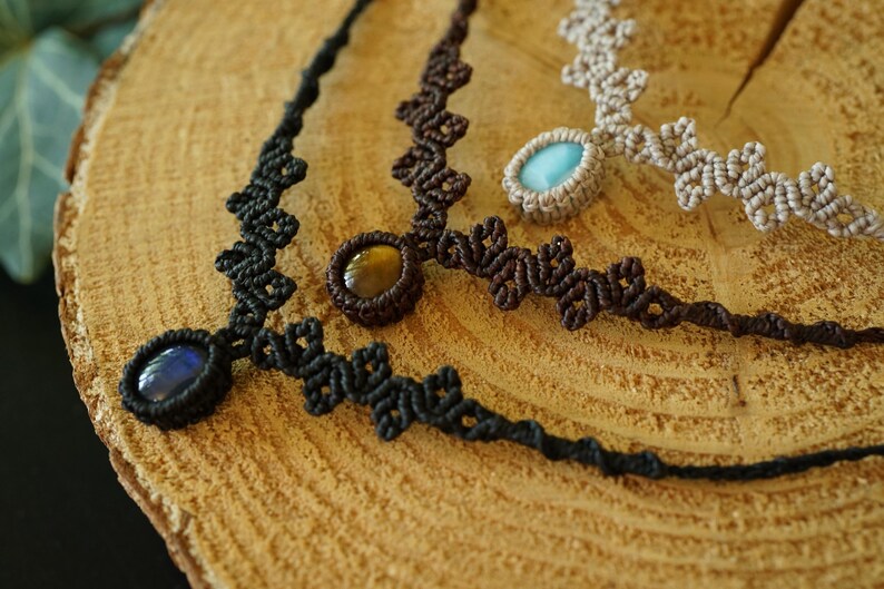 Puede incluir: Tres collares de macram&eacute; con piedras cabuj&oacute;n ovaladas. Un collar es negro con una piedra azul oscuro, otro es marr&oacute;n oscuro con una piedra ojo de tigre marr&oacute;n dorado y otro es beige con una piedra azul claro, todos con nudos intrincados.