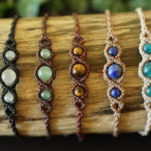 May include: Six handmade macrame bracelets with gemstone beads. The bracelets feature black, brown, and beige cords, with gemstones in white, green, brown, blue, and turquoise. The bracelets are displayed on a piece of wood.