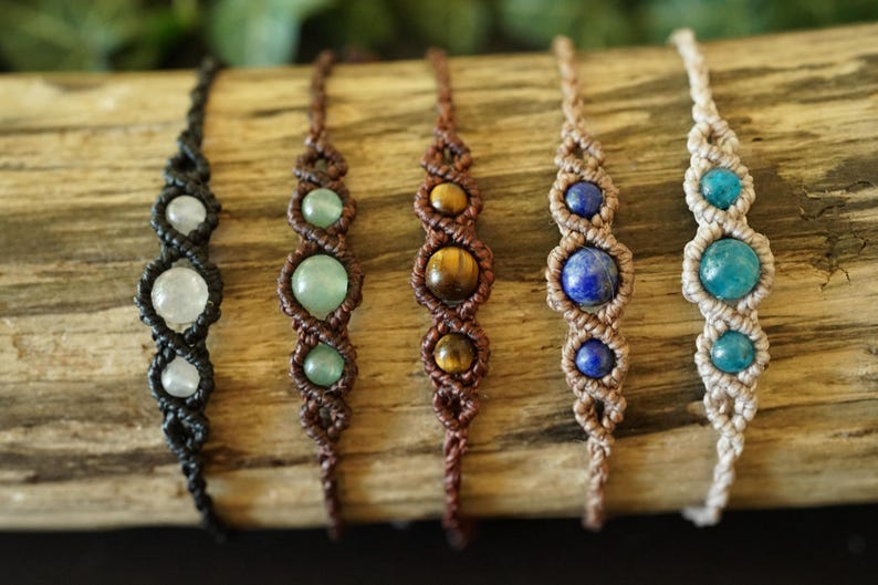 May include: Six handmade macrame bracelets with gemstone beads. The bracelets feature black, brown, and beige cords, with gemstones in white, green, brown, blue, and turquoise. The bracelets are displayed on a piece of wood.