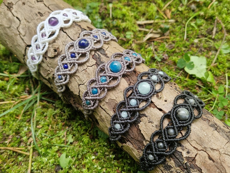 Puede incluir: Cinco pulseras de macram&eacute; con detalles de piedras preciosas se exhiben sobre un tronco de madera. Las pulseras presentan varios colores, incluyendo blanco, gris y negro, con piedras preciosas en tonos p&uacute;rpura, azul y verde. El fondo es un lecho de musgo verde.