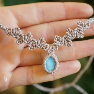 Puede incluir: Un collar de macram&eacute; con una piedra azul claro en forma de l&aacute;grima, engastada en un cord&oacute;n beige anudado. El dise&ntilde;o del collar presenta patrones de nudos sim&eacute;tricos e intrincados que se extienden desde la piedra a lo largo del cord&oacute;n.