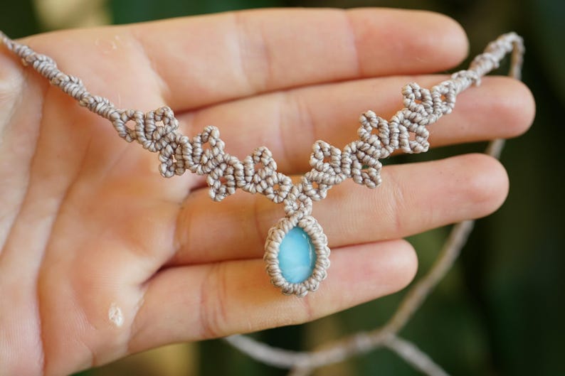 Puede incluir: Un collar de macram&eacute; con una piedra azul claro en forma de l&aacute;grima, engastada en un cord&oacute;n beige anudado. El dise&ntilde;o del collar presenta patrones de nudos sim&eacute;tricos e intrincados que se extienden desde la piedra a lo largo del cord&oacute;n.