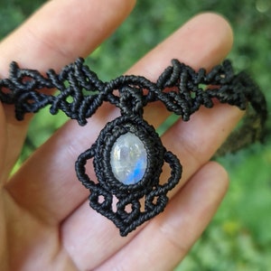 May include: Black macrame choker necklace featuring a central pendant with a pear-shaped, iridescent moonstone. The intricate knotwork forms a decorative pattern around the stone and along the choker's band. A handmade jewelry item.